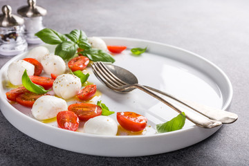 White plate of healthy classic delicious caprese salad with ripe tomatoes and mozzarella cheese with fresh basil leaves on gray concrete background with space for text. Italian food.