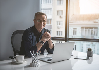 Portrait of cheerful senior businessman noting in laptop while looking at camera. He sitting at desk in office. Profession concept