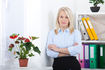 Successful business woman looking confident and smiling. business woman in blue shirt looking friendly into camera standing near window in office