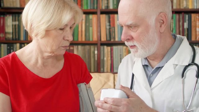 Male professional doctor in white coat with stethoscope at work. Senior man physician measuring pressure to sick senior female patient by tonometer at home. Consulting about treatment and therapy