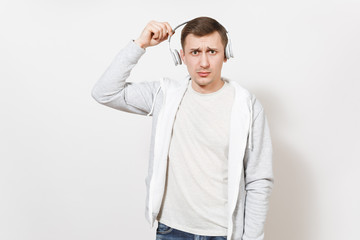 Young handsome student in t-shirt and light sweatshirt listening to music with white wireless headphones and in perplexity removes them in studio on white background. Concept of emotions