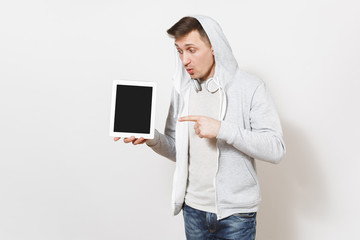 Young handsome surprised student in t-shirt and light sweatshirt with hood with headphones holds a tablet in hands and points to it with finger in studio on white background. Concept of technology