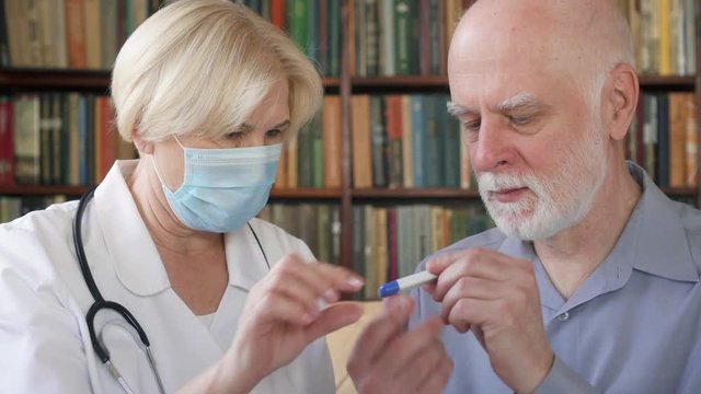 Female professional doctor in medical mask at work. Senior woman physician measuring temperature to sick senior male patient by thermometer at home. Consulting about treatment and therapy