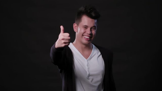 An attractive young man showing a Thumbs Up gesture against a black background. Medium Shot
