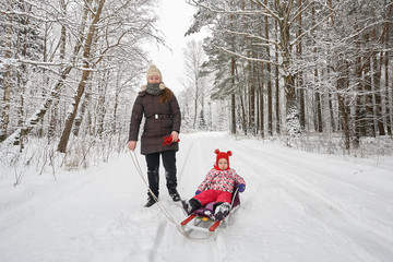 Young beautiful girl whith wihite hat  posing against a winter park background and playing with the girl child with the snow in forest. Towing sledge