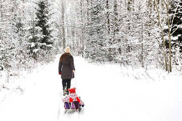 Young beautiful girl whith wihite hat  posing against a winter park background and playing with the girl child with the snow in forest. Towing sledge