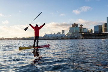 Adventurous man is paddle boarding near Downtown City during a vibrant winter sunrise. Taken in Coal Harbour, Vancouver, British Columbia, Canada.