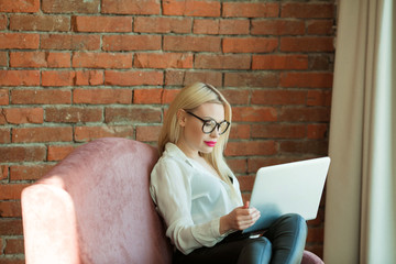 beautiful young girl with blond hair sitting on the couch with a laptop in hand near a brick wall