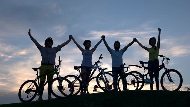 Young cyclists holding hands at sunset. Four young persons with bicycles walking cheerfully together along sunset horizon. Rest and healthy lifestyle.
