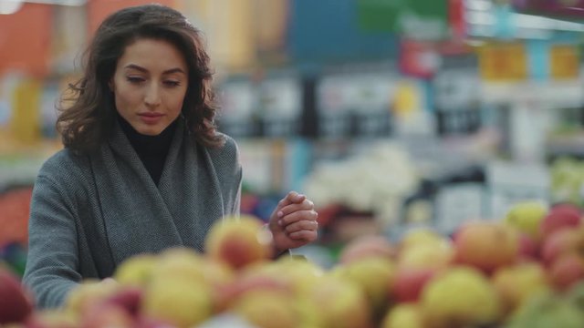 Serious young woman choosing apple at fruit vegetable supermarket marketplace lifestyle commerce product fresh hand customer food beautiful girl shopping selecting face buy store grocery holding