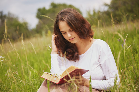 Portrait of lovely beautiful female with attractive look reads book in open air, sits in green field, enjoys summer sunny weather and wonderful nature view. Woman likes calm atmosphere while reading