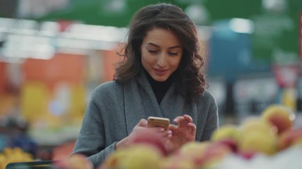 Woman doing grocery shopping at the supermarket and using phone look at camera smile internet technology shopper buying retail shop vegetable market mobile young discount hypermarket online products