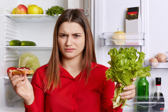 Discontent female frown face, has to keep to diet, eats only fruits and vegetables, holds lettuce and sandwhich in hands, stands at kitchen near refrigerator, has unhappy expression. Food concept