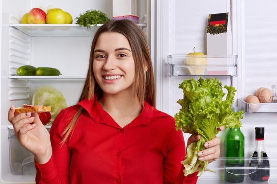 Adorable young woman with happy expression, dressed in red blouse, holds lettuce and sandwhich while stands at kitchen, near opened fridge, going to make delicious breakfast. Positivity and happiness