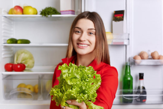 Cheerful beautiful female holds salad lettuce in hands, supports healthy nutrition, stands near fridge. Selective focus. Adorable woman demonstares her favourite product for making vegeterian salad