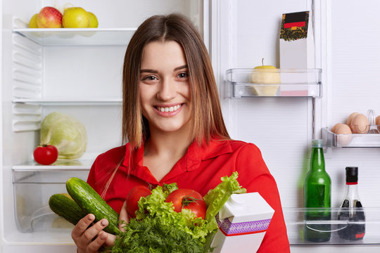 Cheerful lovely young woman with attractive look has broad smile, poses near fridge with fresh vegetables and milk, being in good mood, smiles joyfully, going to prepare delicious breakfast.
