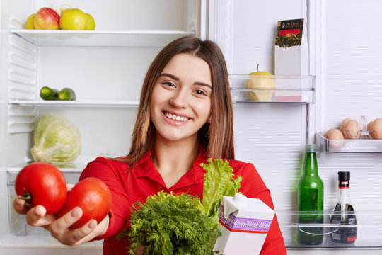 Pretty housewife with delighted look shows products which she bought in grocer`s shop. Beautiful smiling woman holds fresh red tomatoes, lettuce and dill, poses in kitchen against opened fridge