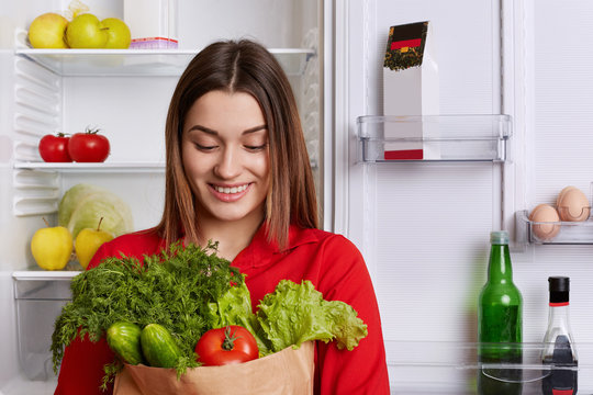 Portrait of happy woman with cheerful expression holds paper bag with tomatoes, cucmbers, dill and lettuce, comes from shop, going to make vegetable salad. People and healthy eatting concept