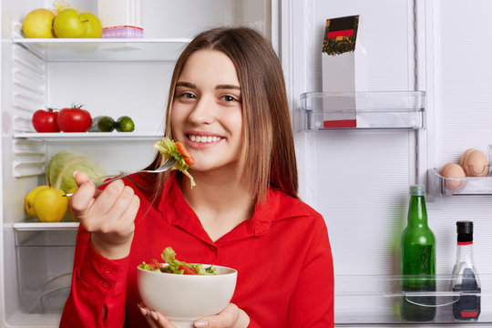 Photo of good looking young woman with pleasant appearance stands near opened refrigerator with bowl of salad, eats only healthy food, being in good mood. People, eatting, vegan and lifestyle
