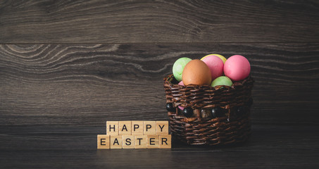 The inscription Happy Easter with light wooden plates and painted eggs in a wicker basket on a dark wooden background