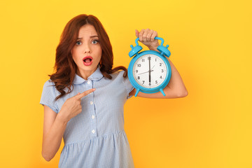 Young woman studio isolated on yellow holding alarm clock pointing at time - obrazy, fototapety, plakaty