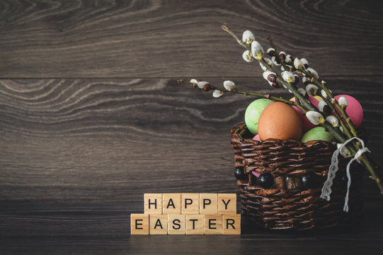 The inscription  happy Easter, made of light wooden cubes, painted eggs in a wicker basket and willow branches with seals on a dark wooden background