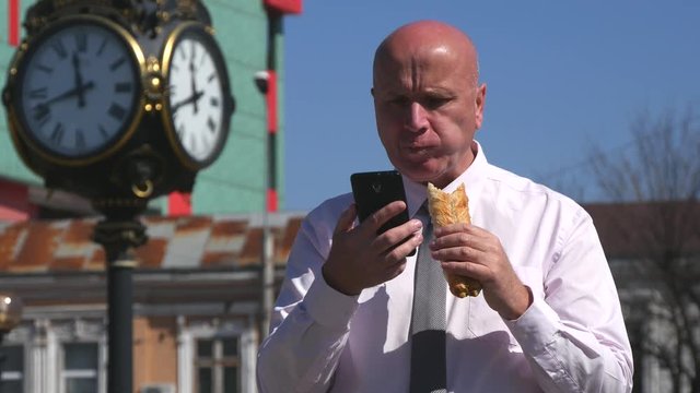 Businessman Outside Office Building Eat a Sandwich and Check Cell Online News
