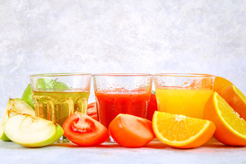 Glasses with fresh orange, apple, tomato juice on a gray concrete table. Lobules Fruits and vegetables around.
