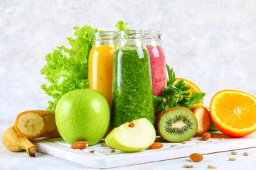 Green, yellow, purple smoothies in currant bottles, parsley, apple, kiwi, orange on a gray table.