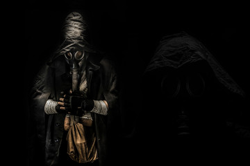 Fototapeta na wymiar man in the gas mask in the hood, on the black background surrounded by smoke, with hands on the chest, survival soldier after apocalypse.