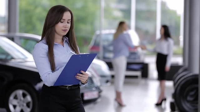 Young Female Car Dealer Writing On Paperboard While In Background Customer Shake Hands With Another One  