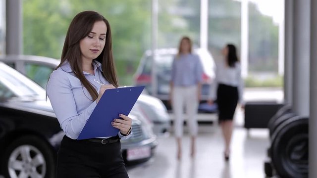 Young Female Car Dealer Writing On Paperboard While In Background Customers Looking For New Car 