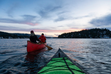 Couple friends on a wooden canoe are paddling in an inlet surrounded by Canadian mountains during a vibrant sunset. Taken in Indian Arm, near Deep Cove, North Vancouver, British Columbia, Canada.