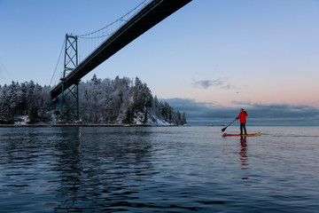 Adventurous man on a Standup Paddle Board is padding near Lions Gate Bridge during a vibrant winter sunrise. Taken in Vancouver, British Columbia, Canada.