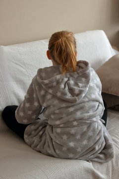 Młoda blondynka siedzi po turecku, samotnie na sofie, wpatruje się w ścianę, ubrana jest  w szary szlafrok i ciemne getry, odwr&oacute;cona tyłem, sofa ma jasną narzutę, jasna ściana w tle