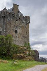Fototapeta na wymiar Dornie, Scotland - June 10, 2012: Gray-brown-stone main house and tower of Eilean Donan Castle. Green vegetation up front. Gray cloudscape and one artillery piece.