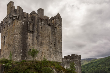 Fototapeta na wymiar Dornie, Scotland - June 10, 2012: Gray-brown-stone main house and tower of Eilean Donan Castle. Green vegetation up front. Gray cloudscape . Mountains as backdrop.