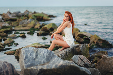 Fototapeta na wymiar pretty redhead woman in white dress posing on rocks at ocean