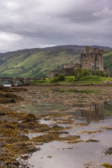 Dornie, Scotland - June 10, 2012: Footbridge, linking green island to mainland, leads to Brown-stone entire Eilean Donan Castle. Under rain-heavy dark cloudscape. Low tide exposes brown seaweed. 