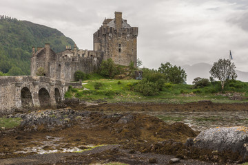 Dornie, Scotland - June 10, 2012: Creek in grass leads to Brown-stone entire Eilean Donan Castle. Under rain-heavy dark cloudscape. Low tide exposes brown seaweed. Green mountains.