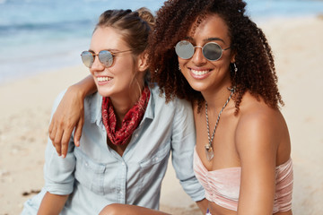 Adorable woman with Afro hairstyle embraces her female partner, beinng lesbains, enjoy togetherness and calm atmospere at seaside, admire views, have happy looks. People, lifestyle, recreation