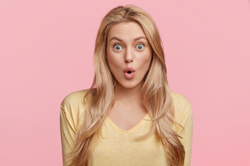 Isolated shot of pretty young female stares at camera with shocked expression, recieves unexpected news, keeps lips rounded, poses in studio over pink blank wall. Great surprisment or unexpectedness