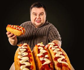 Fat man eating fast food hot dog on plate. Breakfast for overweight person. Junk meal leads to obesity. Person regularly overeats concept on black background. Health problems due to malnutrition.