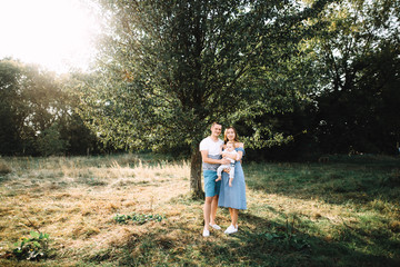 Naklejka na ściany i meble Happy family. Parents with their little son stand near a big tree in the nature at sunset