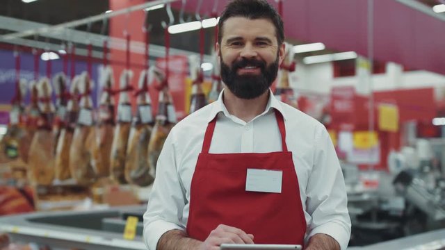 Portrait seller man with beard in supermarket look camera smile in the meat department happy business beautiful fresh produce retail shop worker food choosing young grocery model shopping