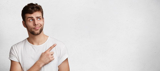 Puzzled blue eyed handsome young bearded man poses agianst blank copy space, dressed casaully,...