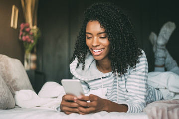 close up of a pretty black woman with curly hair smiling and using phone on bed looking away