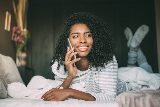 close up of a pretty black woman with curly hair smiling and using phone on bed looking away