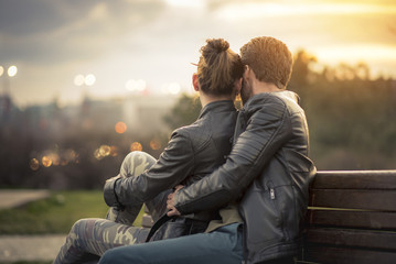 Young sweet couple embraced on a bench in park watching a beautiful sunset, enjoying their love and...