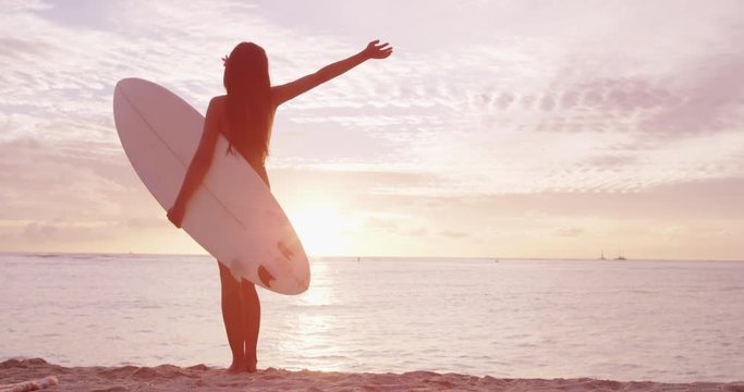 Surfer girl surfing happy and serene looking at beach sunset holding surfboard. Female surfer woman looking at water with arms outstretched living healthy active lifestyle. SLOW MOTION shackycam